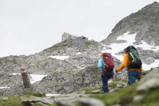 Zwei Wanderer im Hochgebirge, im Hintergrund eine Alpenvereinshütte auf einem kleinen Felsplateau.