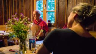 Mensch sitzen um Tisch in Hütte, ein Mann spielt Akkordeon