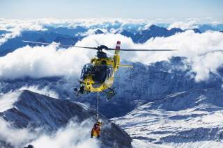 Ein gelber Hubschrauber fliegt durch die verschneiten Alpen, ein einem Rettungsseil hängt ein Einsatzhelfer.