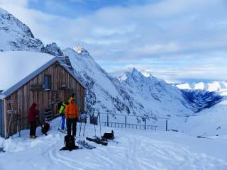 Menschen mit Ski stehen vor kleiner Hütte in winterlichem Gebirge