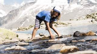 Kind steht im Gebirgsfluss und greift nach Steinen, schaut in die Kamera. Im Hintergrund ragen die Gipfel in den Himmel.