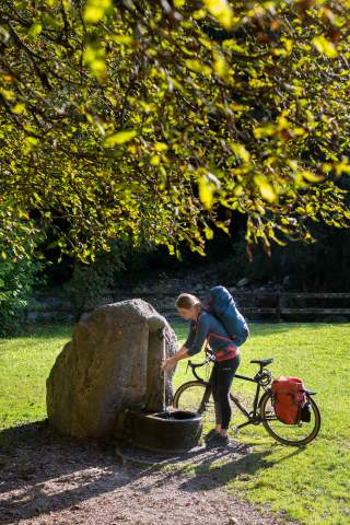 Lena Müller steht mit ihrem Fahrrad unter einem Baum und kühlt ihre Hände an einem Brunnen ab.