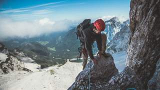 Mann mit Kletterausrüstung steigt auf einen Felsvorsprung. Im Hintergrund ergibt sich der Blick ins Tal.