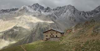 Schutzhütte aus braunen Steinen, im Hintergrund graubraune Gipfel die leicht mit Schnee bedeckt sind.