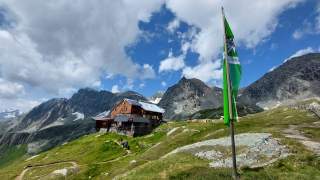 Eine Alpenvereinshütte im Hochgebirge. Im Vordergrund eine grün-weiße Alpenvereins-Flagge.