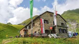 Vor einer Alpenvereinshütte im Hochgebirge wehen mehrere Fahnen; Menschen sitzen auf der Terrasse.