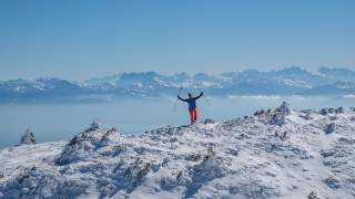 Mann reckt die Hände in die Luft und steht auf einem Grat. Im Hintergrund sieht man die Alpenkette, schneebedeckt.