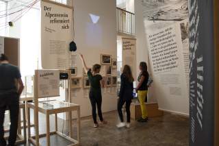 Junge Frau zeigt Besucherinnen bei einer Führung durch das Alpine Museum eine Schautafel