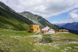Eine große Alpenvereinshütte steht auf einem Wiesenplateau im Hochgebirge. Ganz klein: Zwei Wanderer laufen zur Hütte.