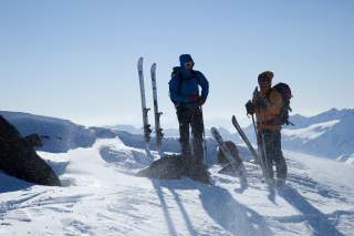 Zwei Menschen mit Tourenski stehen bei Sonne auf windigem Gipfel