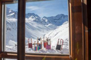 Blick aus einem Hüttenfenster, davor sechs Rodel vor winterlich verschneitem Hochgebirge