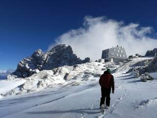 Mensch läuft im Schnee auf futuristisch anmutende Berghütte zu