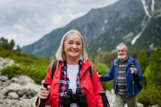 Ein älteres Paar ist beim Wandern in den Bergen unterwegs. Sie strahlend im Vordergrund mit Fernglas um den Hals hängend und in roter Jacke.