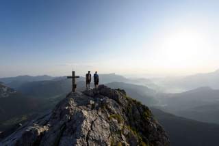 Zwei Menschen stehen neben Gipfelkreuz und genießen die Aussicht