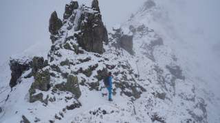Frau auf dem schmalen Pfad Richung Gipfel. Man sieht, dass er sehr schmal ist. Es liegt Schnee. Die Felsen ragen schroff in die Höhe.