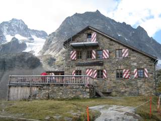 Eine Berghütte mit rot-weiß-gestreiften Fensterläden und dicken Steinwänden in hochalpiner Landschaft.