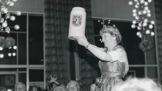 Ein Schwarz-Weiß-Bild zeigt eine Frau in einem Festsaal. Sie trägt ein Dirndl und hebt einen großen Steinkrug.