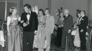 Ballgesellschaft. Ein altes Schwarz-Weiß-Foto zeigt vier Tanzpaare in festlicher Kleidung. Sie laufen in Zweierreihen an gedeckten Tischen vorbei aufs Parkett.