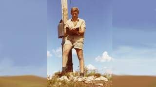Josef Klenner steht am Gipfelkreuz. Ein altes Farbfoto eines jungen Mannes, der an einem Gipfelkreuz lehnt.. Er trägt Lederschuhe, beige Shorts und ein kurzärmeliges Hemd. In der Hand hält er ein Gipfelbuch.
