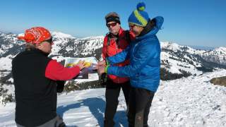 Am Gipfel des Riedberger Horns stehen drei Männer in Winterkleidung. Sie deuten auf eine Illustration, die die geplanten Erschließungen am Berg darstellen. Die Landschaft ist verschneit.