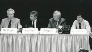 Schwarz-Weiß-Foto von vier Männern. Sie sitzen an einem Tisch mit Husse, vor ihnen stehen Namensschilder.