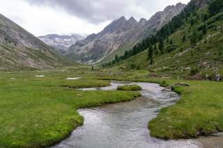 Frei fliessender Bach schlängelt sich durch grüne Moorflächen in Tal umgeben von felsigen Gipfeln