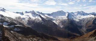 Eine Bergkette mit Gletschern ist leicht verschneit, während die Landschaft Richtung Tal grau-braun ist.